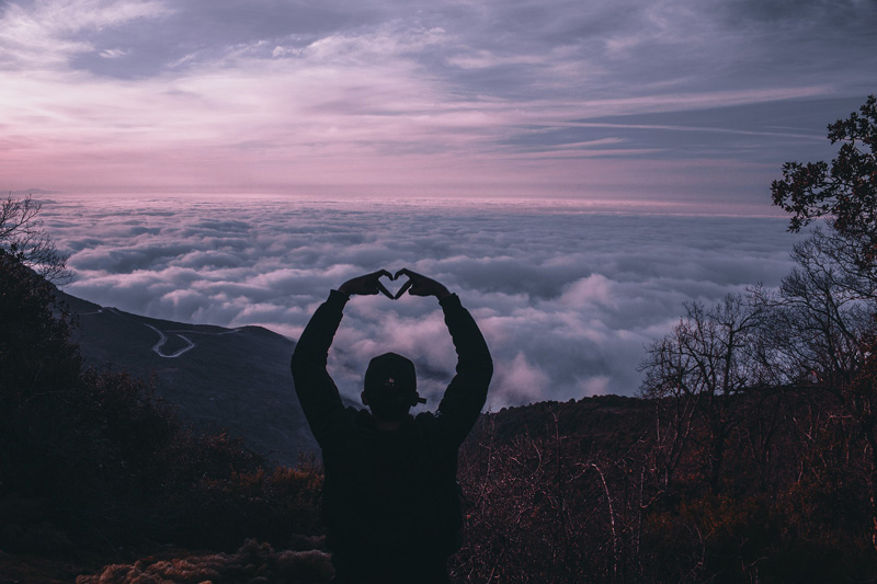 Person sitting atop mountain at dusk looking down on clouds and making heart shape with hands