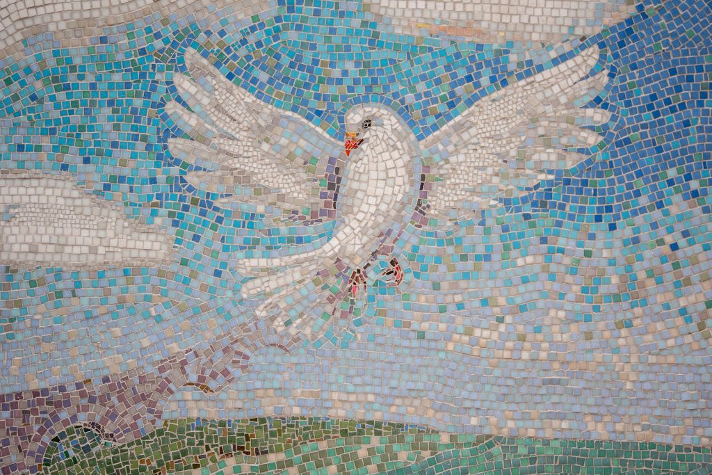 Mosaic artwork of white dove in flight against bright blue sky above green landscape with aqueduct bridge