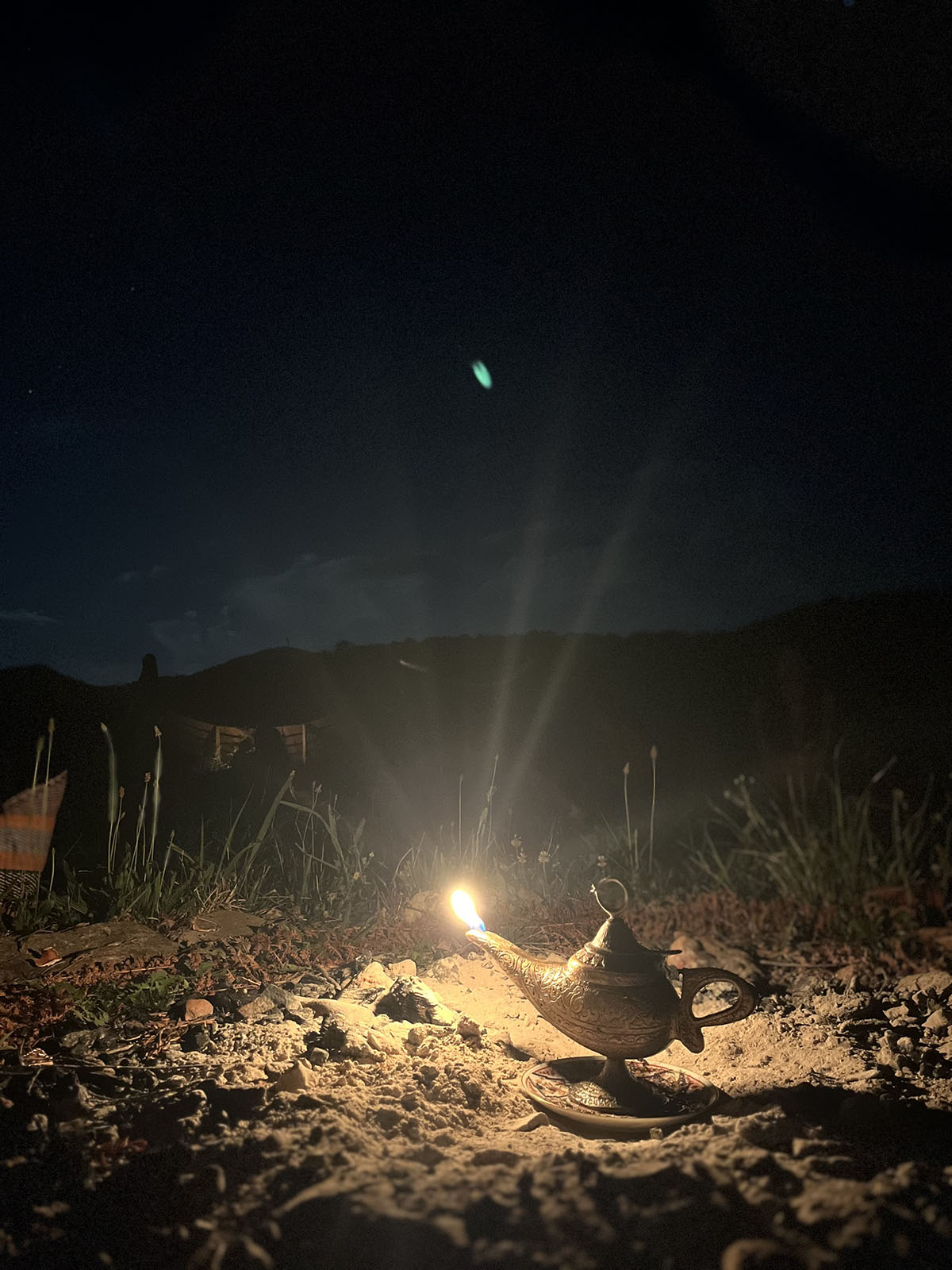 A persion style oil lamp oon the ground at night with a green comet trail flaring in the sky